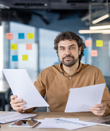 Hombre con camisa marrón sosteniendo dos hojas de papel, con notas adhesivas de colores al fondo